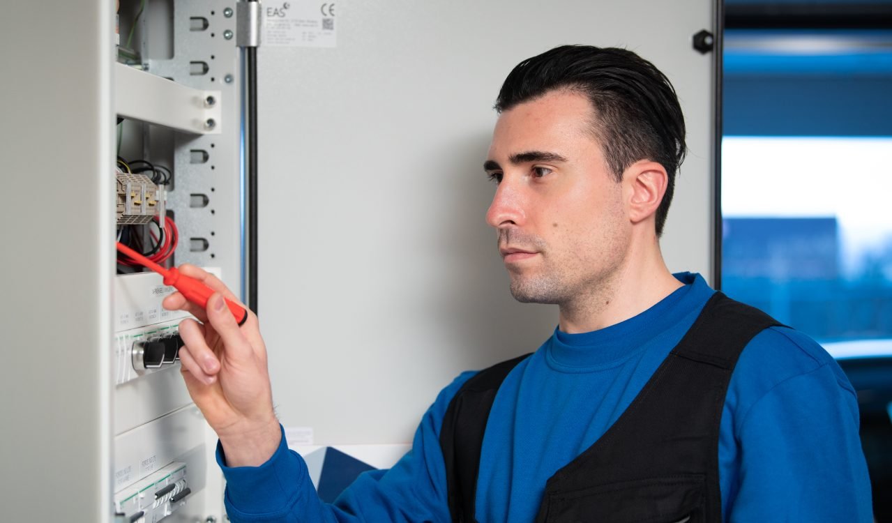 En elektriker i gjenkjennelig Norgeseliten-blå genser monterer strømbryteren i sikringsskapet.